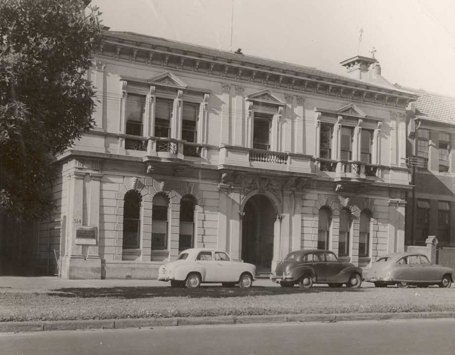 black and white photo of two-storey 19th-century building with three mid- 20th century cars parked in front of it. - click to view larger image