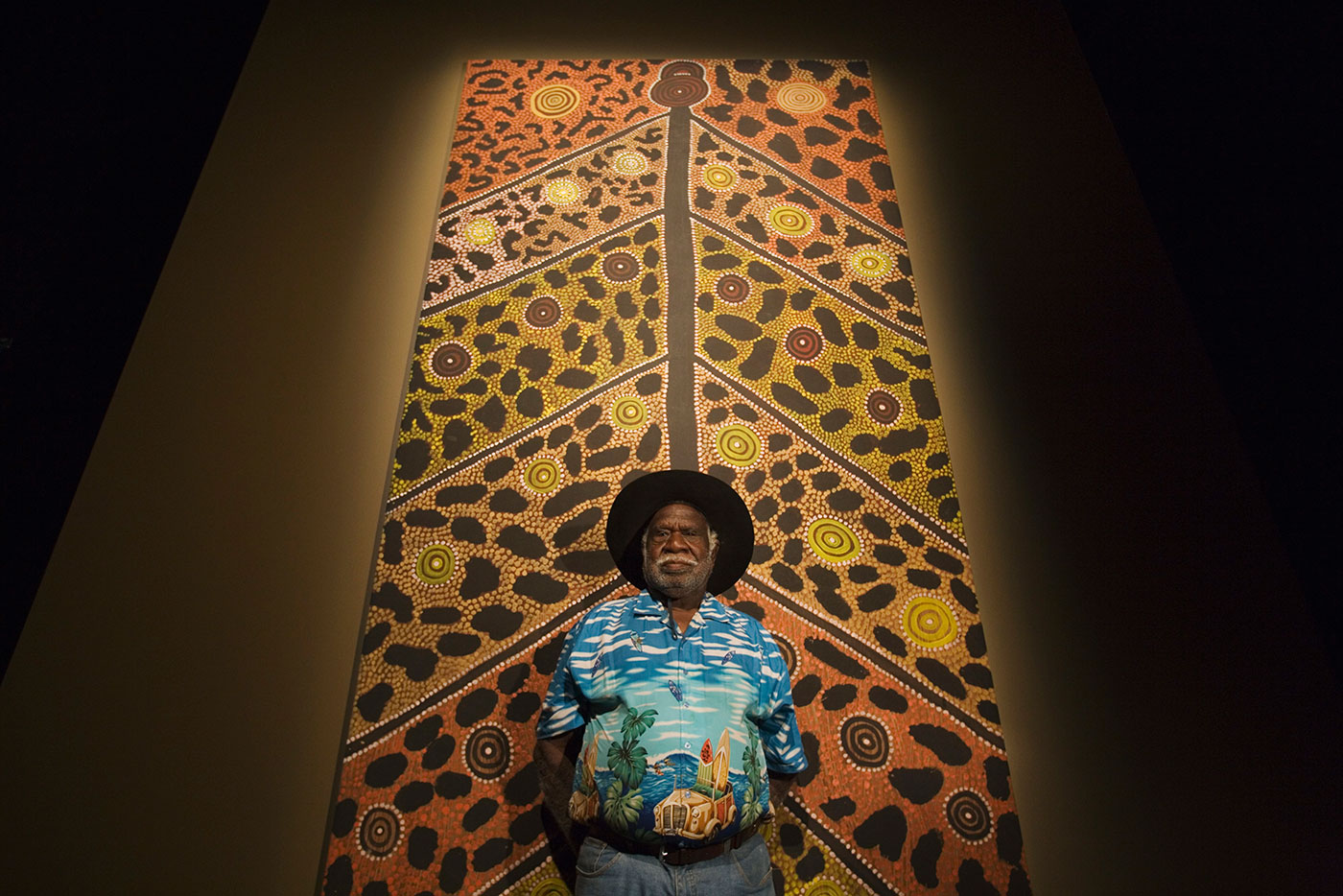Long Jack Phillipus Tjakamarra standing in front of large canvas. - click to view larger image