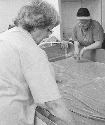 A black and white photo featuring people leaning against a giant-sized sink soaking a very large piece of material.