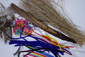 A collection of craft materials are laid out on a white background. Some materials are plant-based and have the appearance of wood and brown string-like material, and others are a mixed coloured ball of wool and a pile of blue, black, white, orange and yellow pipe-cleaners. 