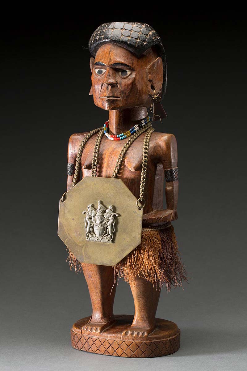 A carved wooden female figure from Papua New Guinea with a plant fibre skirt. The figure is wearing a necklace of blue, turquoise, red, white and mustard coloured beads. A metal chain is draped around the neck of the figure with an African Chief's badge decorated with a silver coat of arms from Rhodesia. A section on the base is separate from the rest of the carving. - click to view larger image