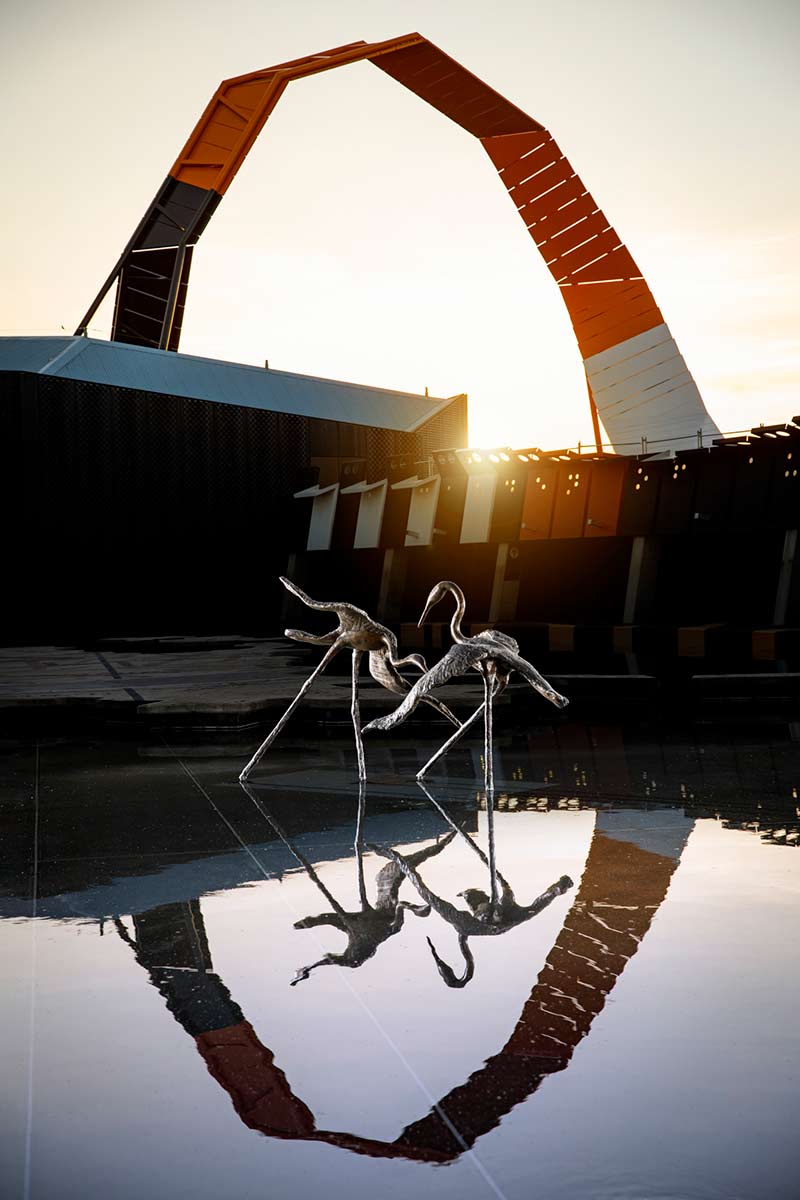 A sunset photo featuring a bronze sculpture consisting of two separate brolga figures. The display is set in a large body of water with the National Museum of Australia building and its bright swooping loop behind it which is reflected in the water. - click to view larger image