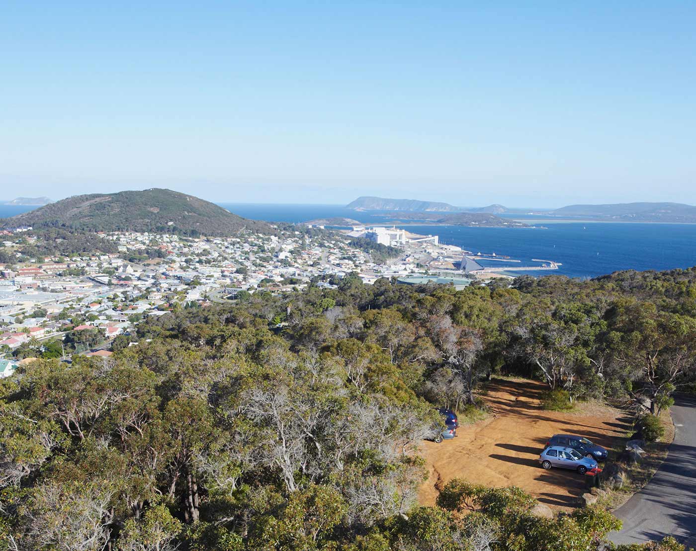 A photo looking down on a dirt carpark with three vehicles visible, surrounded by hundreds of trees. The sea, hills and town form the backdrop.