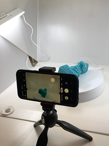 A photograph of a mini tripod and mobile phone positioned for photographing a small blue object inside a lightbox. 