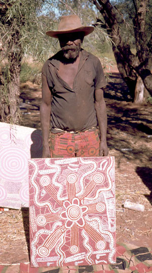 Portrait photo of an Aboriginal Australian man wearing a brown T-shirt and hat andholding a painted canvas. - click to view larger image