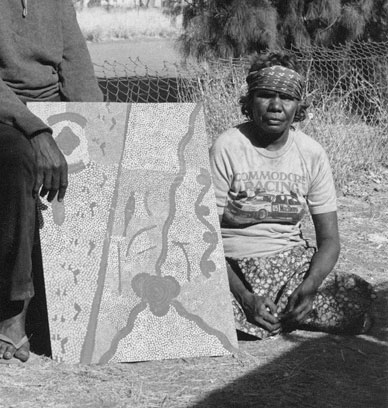 Portrait photo of an Aboriginal Australian woman sitting on the ground alongside a painting. - click to view larger image