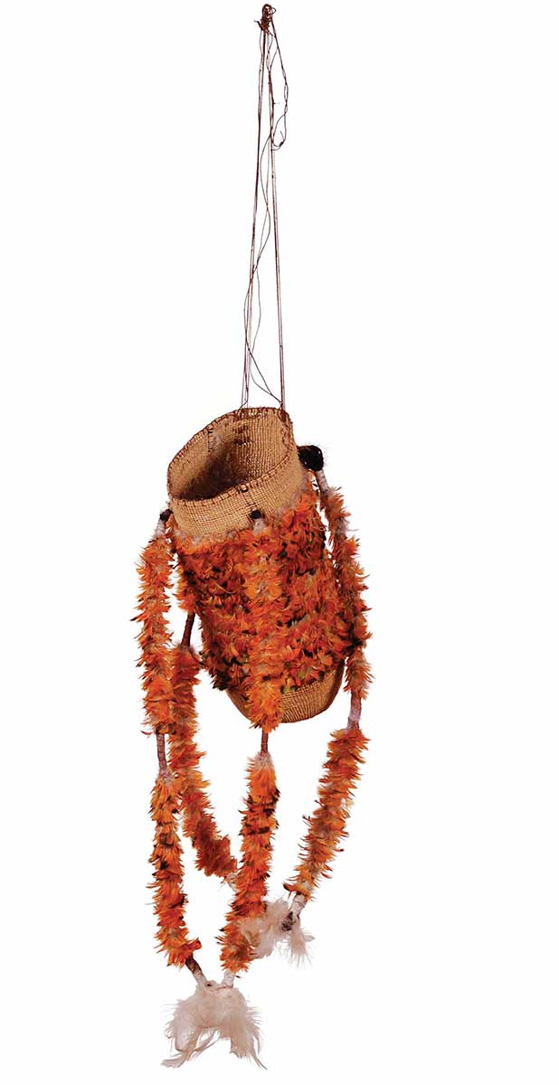 A basket made by an Indigenous Australian. The basket is cylindrical and made from natural fibres. It has a long string handle attached to one side of its opening. Four thick string tassels hang from the basket. Each is decorated with orange and white bird feathers. The basket has a broad band of orange feather decoration around it.