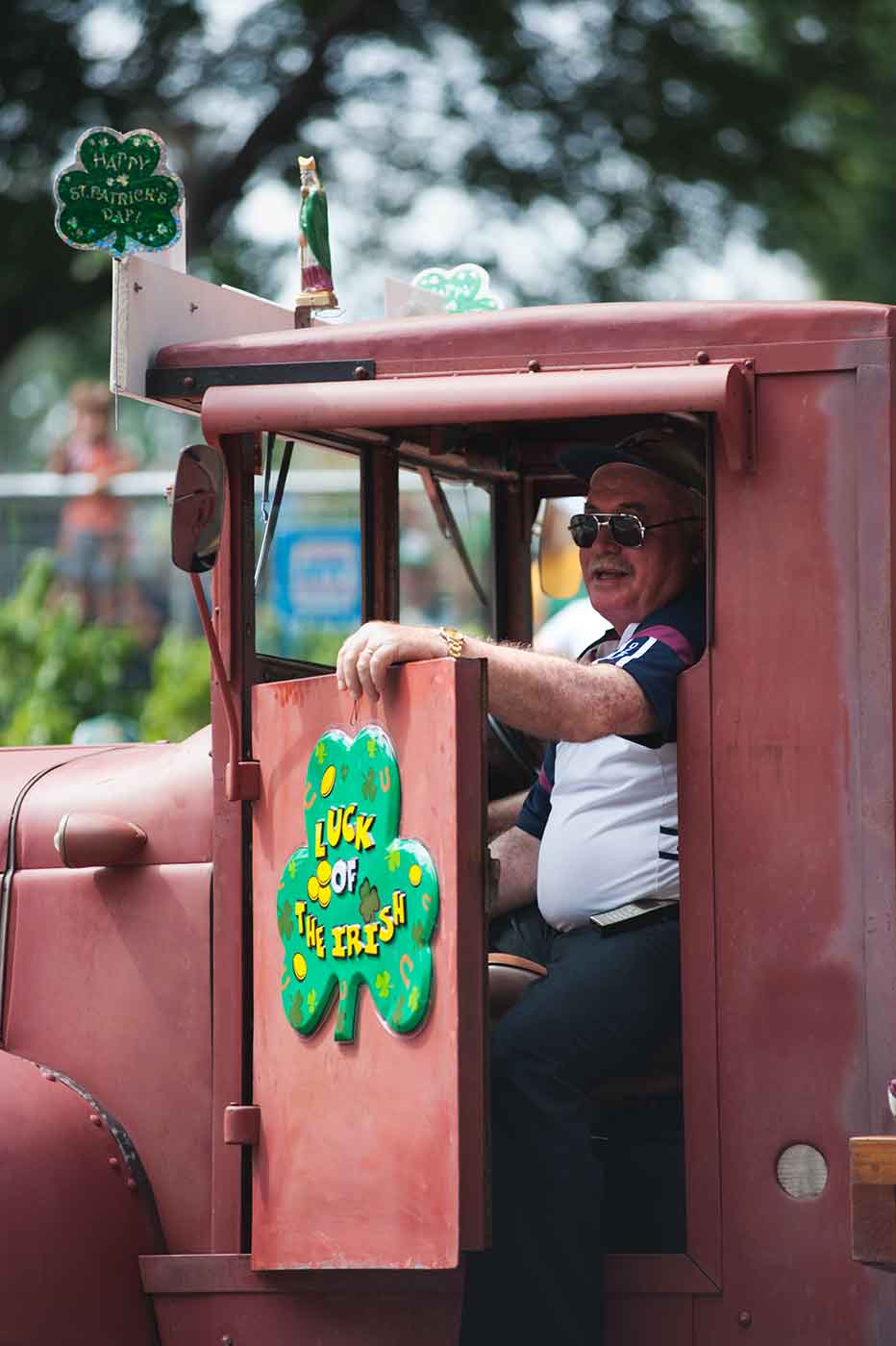 A man sits in a stationary red truck with a sign fixed to the car door of a clover and the text: LUCK OF THE IRISH. - click to view larger image