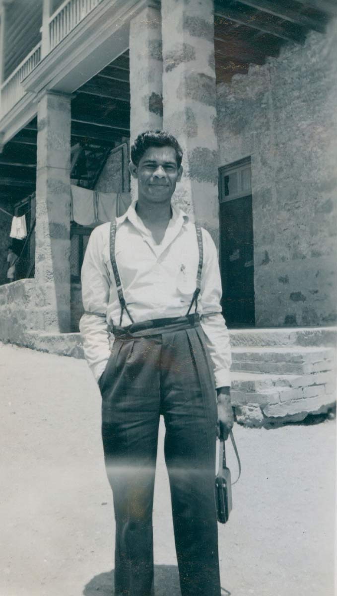 Black and white portrait photo of a First Australian young man wearing slacks and standing in front of the entrance to a sandstone building. - click to view larger image