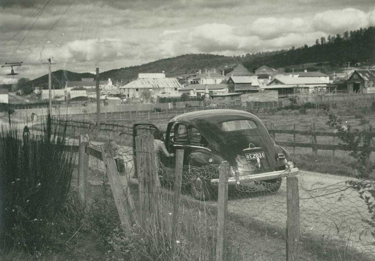 Black and white photo of a car parked on the side of a dirt road heading in the direction of a town which can be seen in the distance.