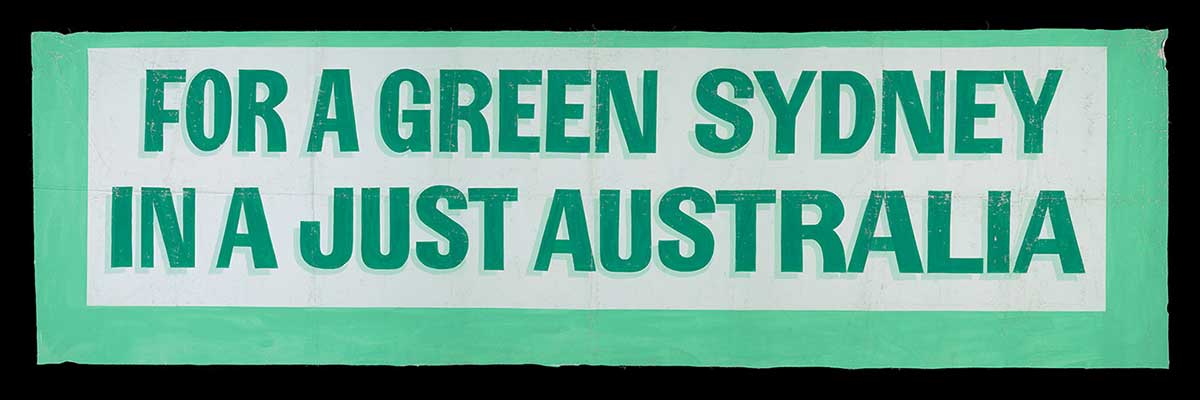 A rectangular banner painted white with a green boarder. It has dark green text with a light green shadow that reads 'FOR A GREEN SYDNEY / IN A JUST AUSTRALIA'.
