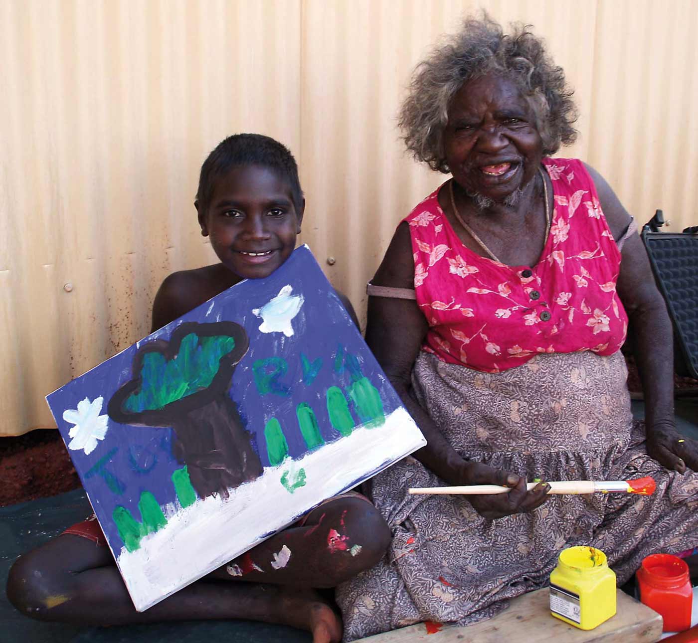 Colour photograph of an elderly woman sitting next to a young boy who is holding a painting. - click to view larger image