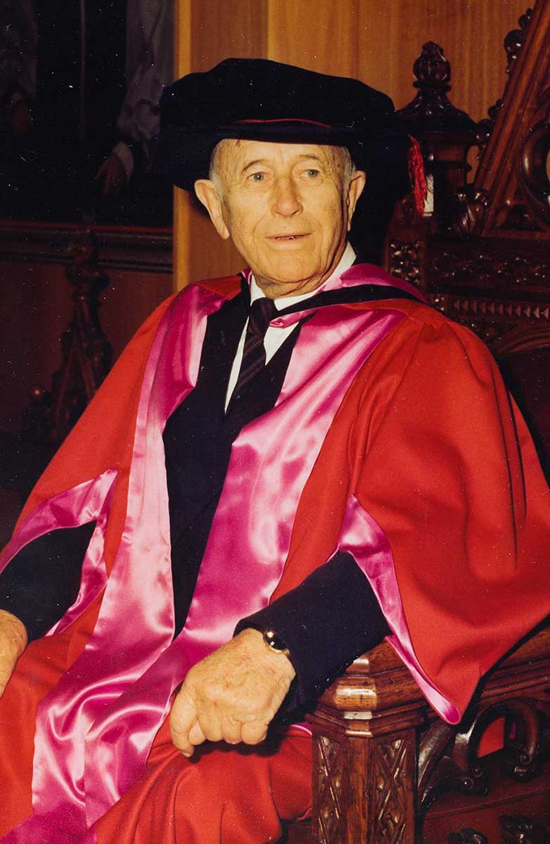 An elderly man wearing university ceremonial clothing is sitting in an ornate wooden chair. - click to view larger image