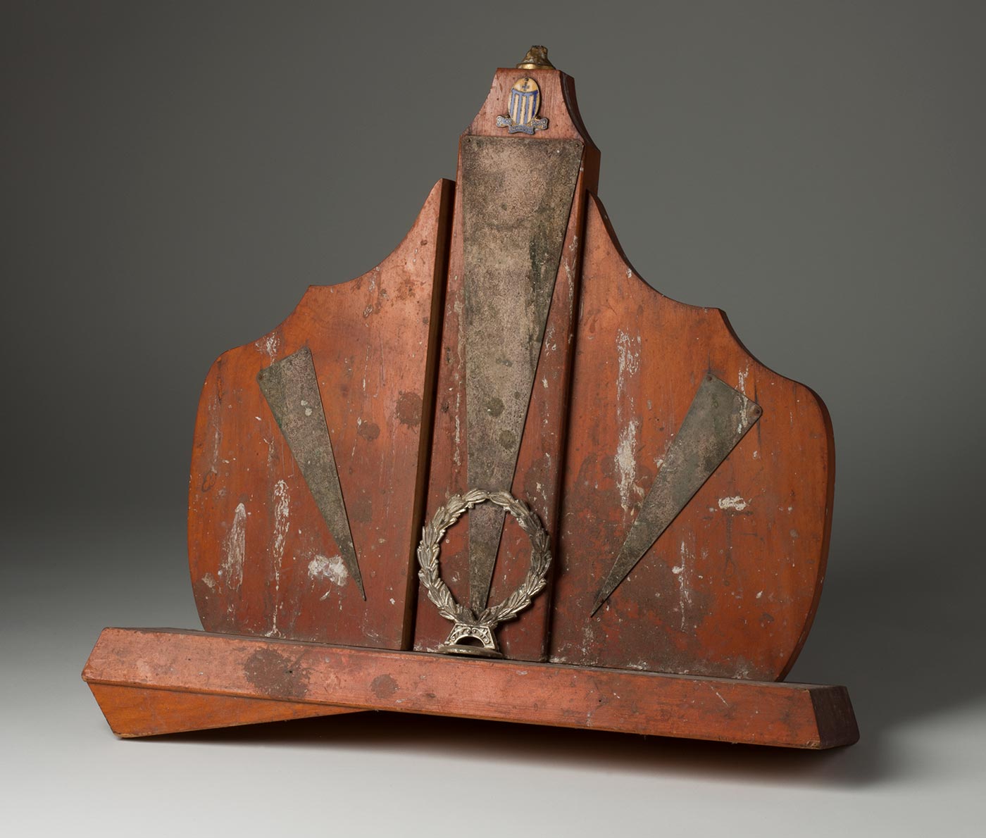 Wooden and metal trophy in a semi-shield shape with a laurel wreath in the centre. At the top there is a blue and yellow striped enamel badge with the words 'O.L.M. PREPARATORY SCHOOL' Remnants of bird droppings streak the trophy. - click to view larger image