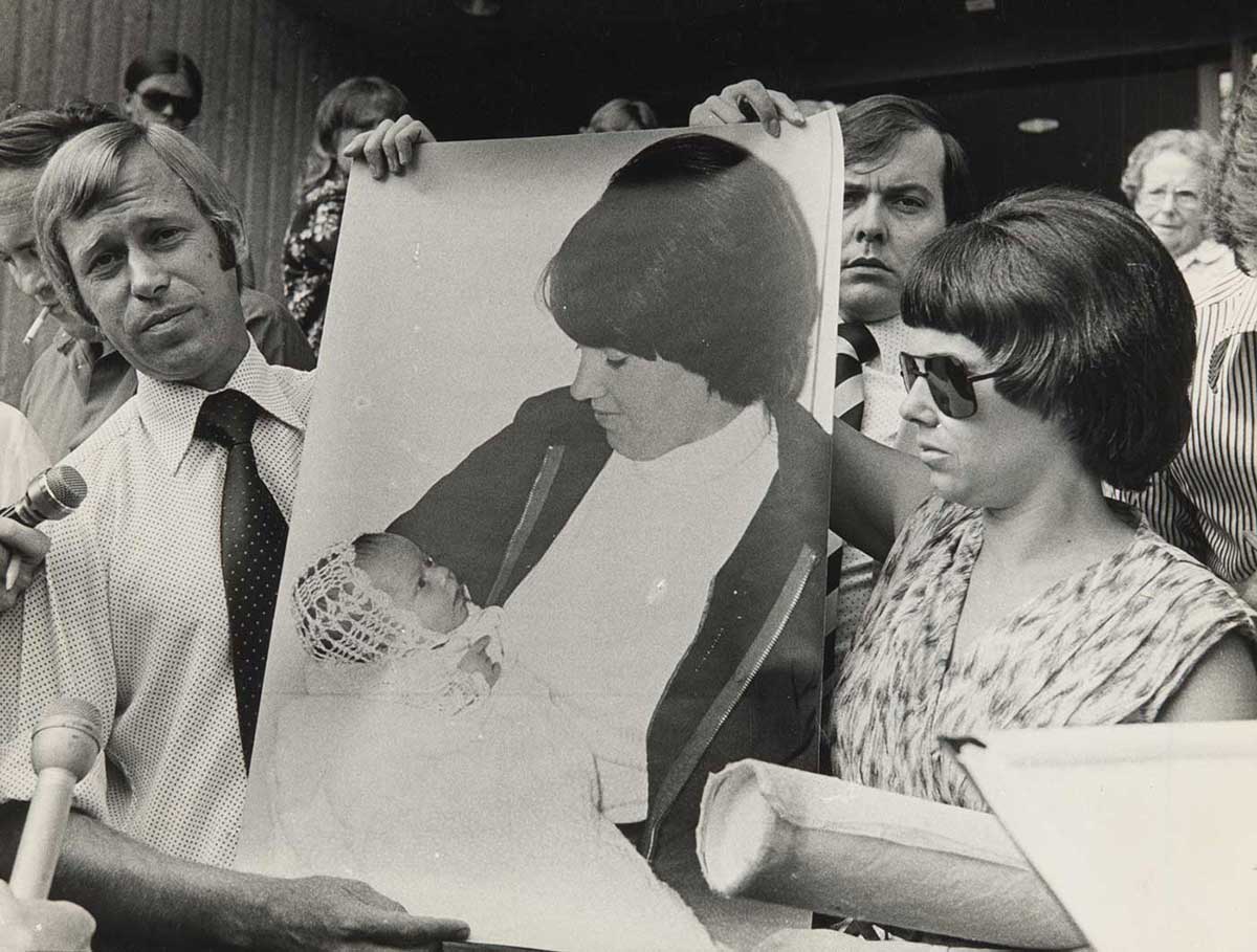 A man and a woman surrounded by the media are holding up a poster-sized print of a photograph of the woman holding a baby.