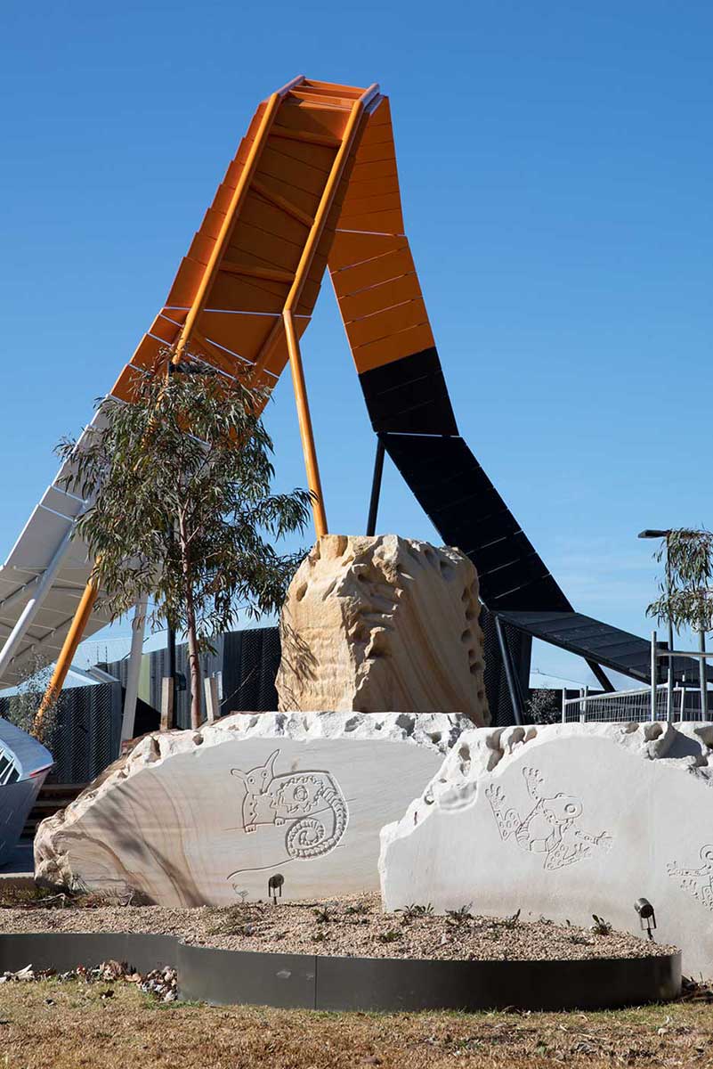 View of a large sculptural architectural feature rising above landscaped grounds of native plants. In the foreground there are large stones with etchings of Australian wildlife. - click to view larger image