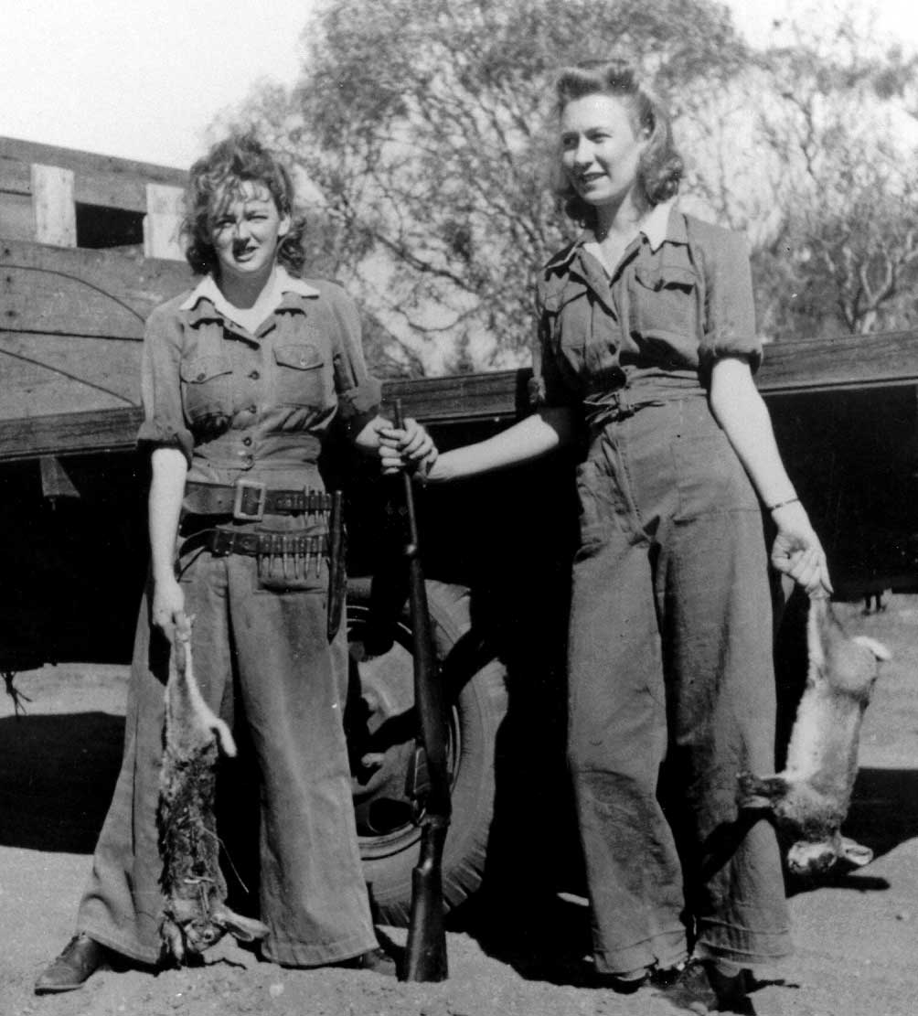 Black and white photo showing two women standing beside the tray of a truck. Both hold a dead rabbit by the hind legs and rest their hands on a rifle at centre. - click to view larger image