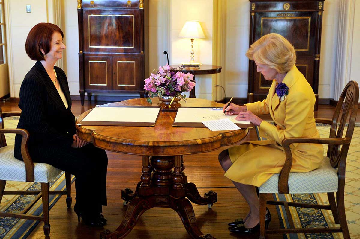 Julia Gillard is sitting at a table with Quentin Bryce who is signing an official document.