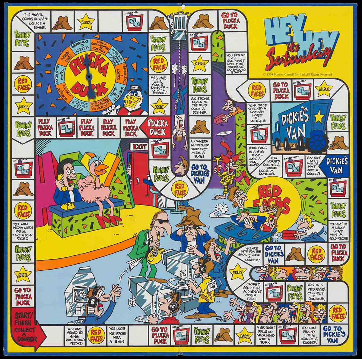 A game board with blue paper exterior and game surface interior, inscribed 'HEY HEY it's Saturday'. The interior shows white game spaces, text and illustrations in colour. A blue plastic arrow is affixed to the board in the upper left quadrant. - click to view larger image