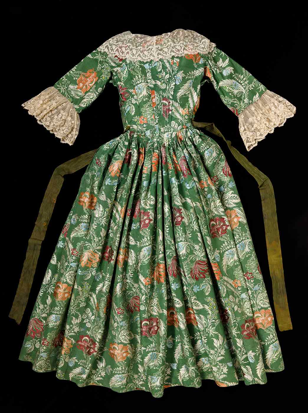 The back of a green and floral dress with lace trim on the sleeves and collar with a long green sash like belt stitched at the waist. - click to view larger image