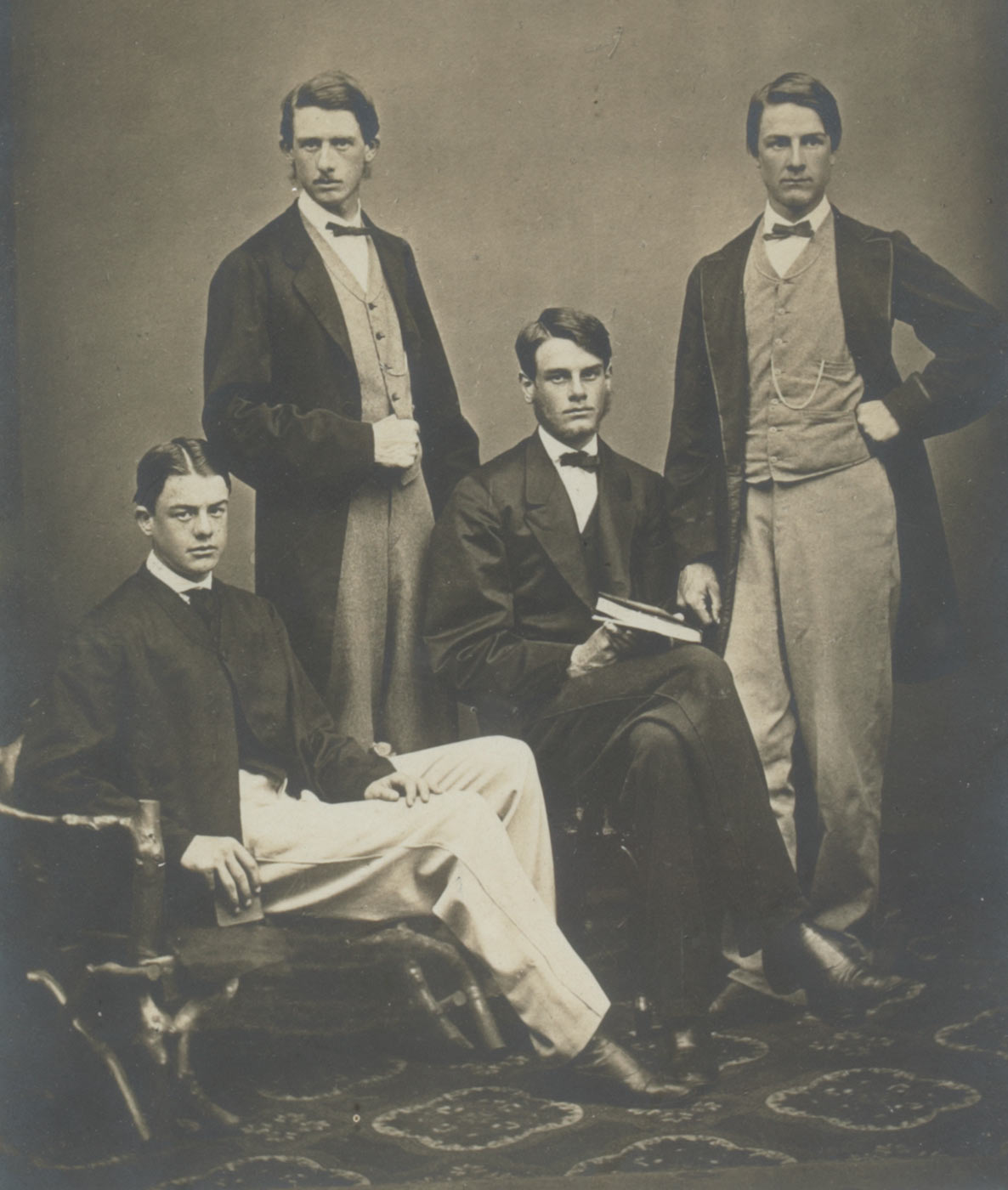 A black and white portrait photograph of four men, two seated and two standing, all wearing suits and bowties. The photograph is set on a grey mount and in a light brown wooden frame with recessed gold interior edging. It depicts standing, on left, William Percy, on right, George Ernest; seated on left, Reginald, on right Henry Montague. Handwritten on mount 