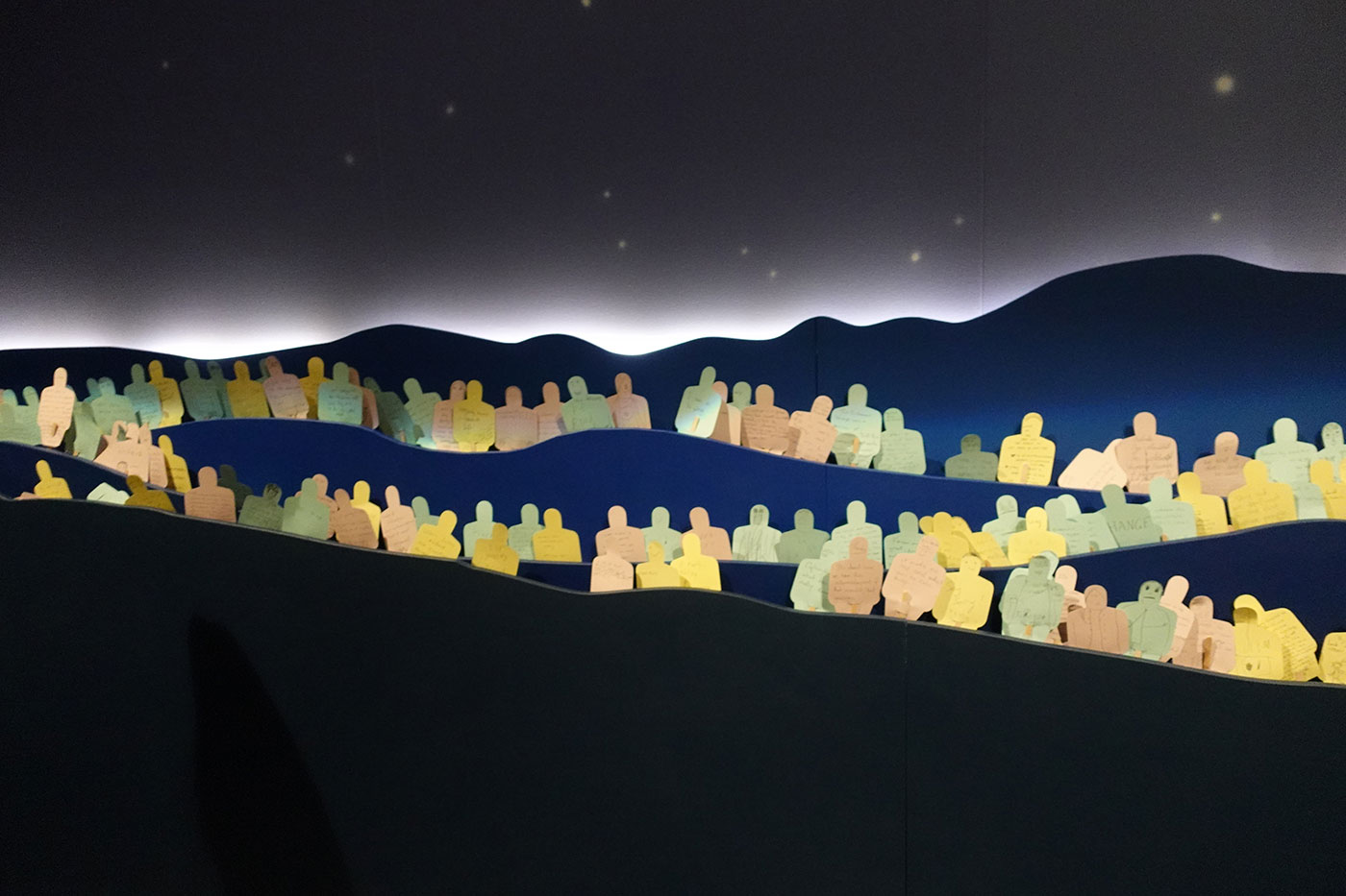 Exhibition display featuring multiple pieces of coloured paper cut out in the shape of people with hand written messages. They are positioned amongst a stylised landscape of mountains and a star-lit sky. - click to view larger image