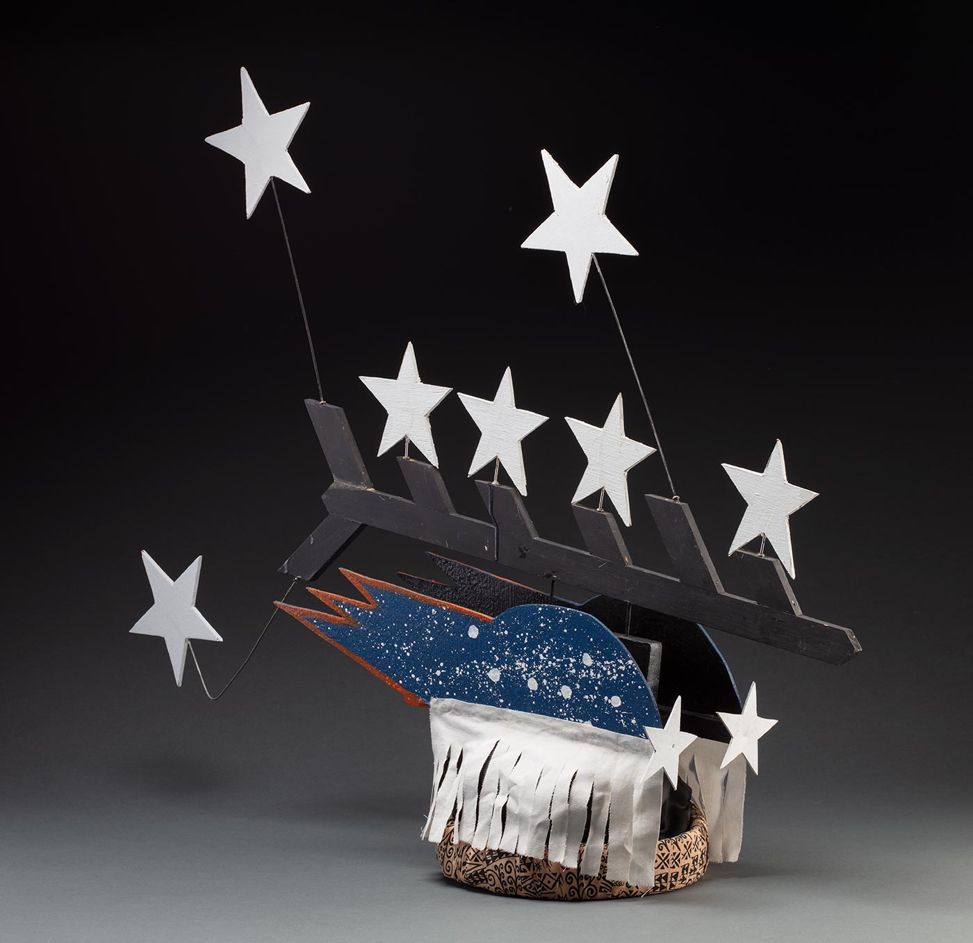 A headdress featuring seven stars protruding from a black painted piece of board forming half a fishbone pattern. It is connected to two pieces of board painted blue with white splatter and seven stars on each side with a white fabric fringe at the bottom. It is all attached to a fabric wrapped headband in the centre. - click to view larger image