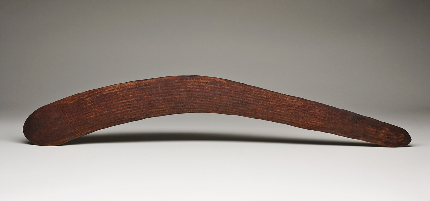 An asymmetrical dark brown wooden boomerang with incised decoration on both sides. The boomerang is slightly convex on one surface and almost flat on the other. One end is semicircular while the other is a narrower rounded point. Both sides have three sections of parallel horizontal grooves. On the convex surface there are three grooves at the larger end, seven grooves at the centre and two grooves at the narrower end. On the flat surface there are three curved grooves at the larger end, six grooves at the centre and three grooves at the narrow end. - click to view larger image