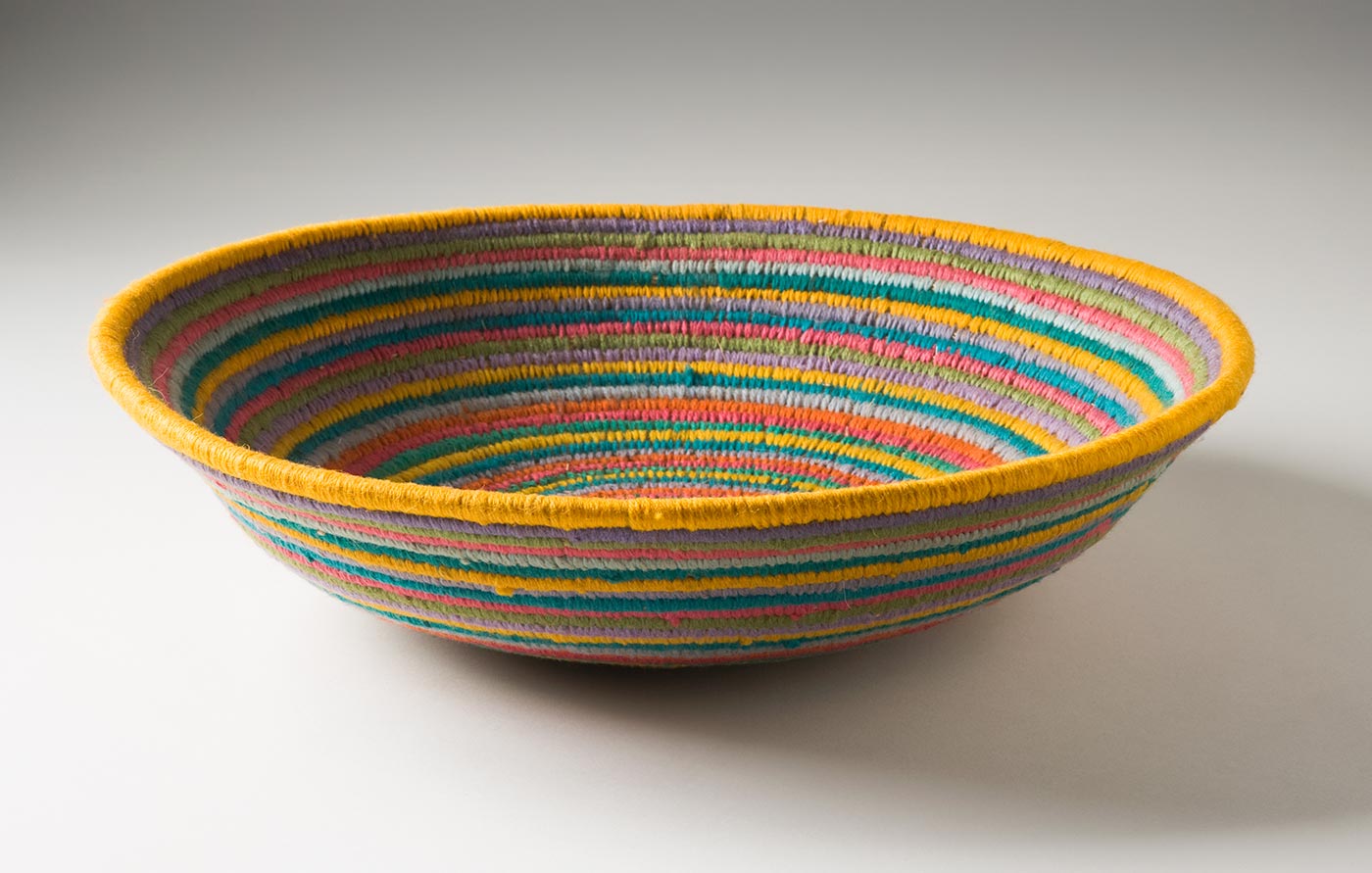 A multi-coloured circular coiled bowl-shaped basket made of yarn and plant fibre. The centre of the basket is in yellow yarn followed by horizontal stripes of yarn in lime green, lavender, pink, orange, yellow and light blue grey. - click to view larger image