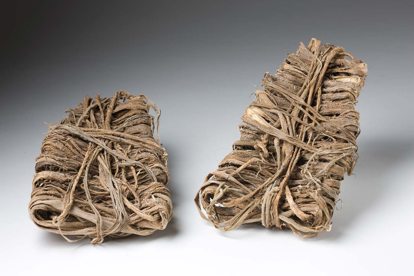 A pair of sandals made of plant fibre. They have a trapezoid shape and are made from strips of bark wrapped around and layered on top of each other. - click to view larger image