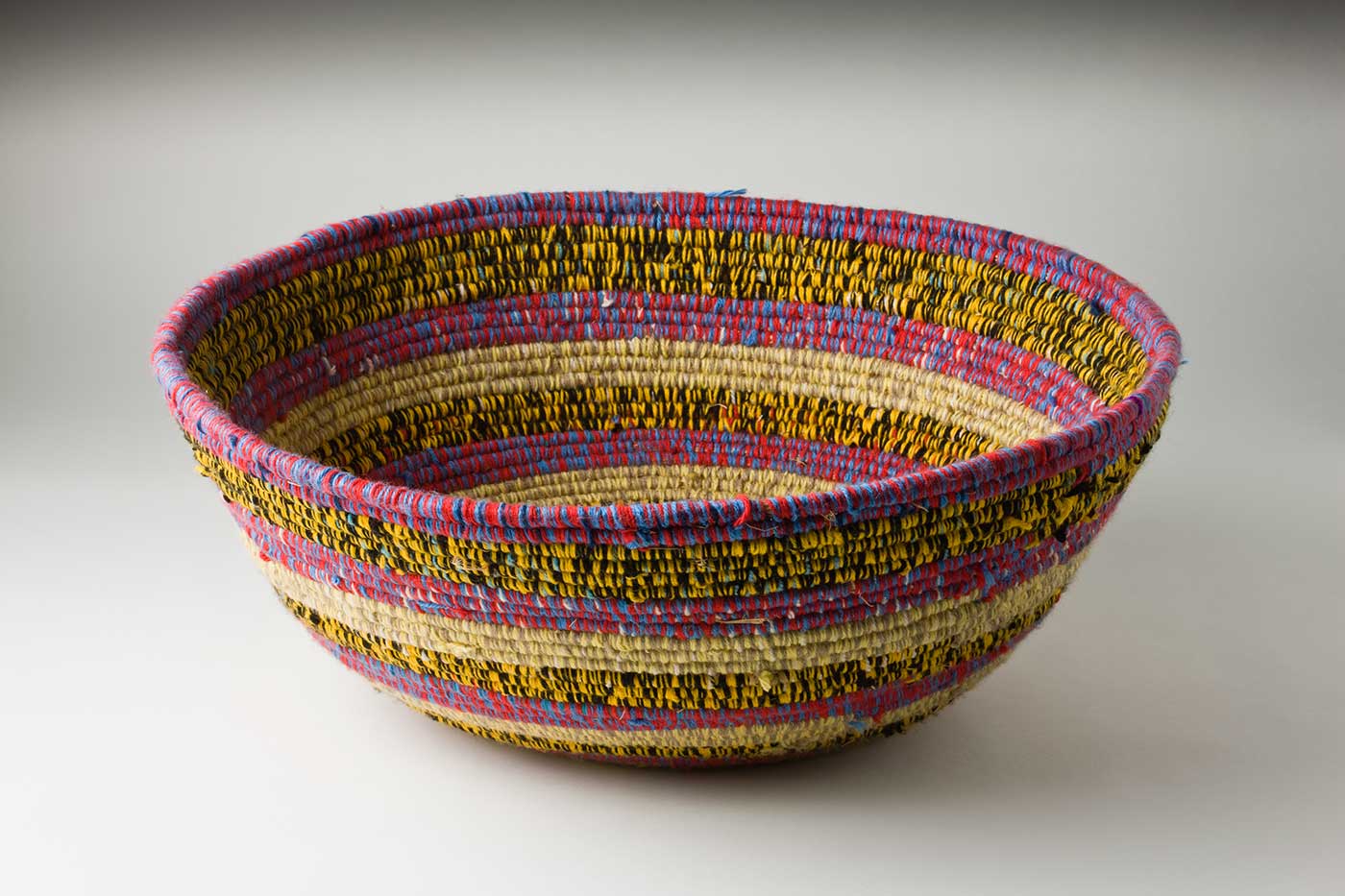 An oval coiled multicoloured yarn and plant fibre basket. The plant fibre in the centre of the basket is covered by purple-yellow yarn, followed by horizontal alternate yarn coverings over the fibre. The yarn colours are green-yellow, tan-yellow, purple-red, yellow-black, green-silver-brown and red-white-brown. The top plant fibre is covered by yellow-black yarn. - click to view larger image