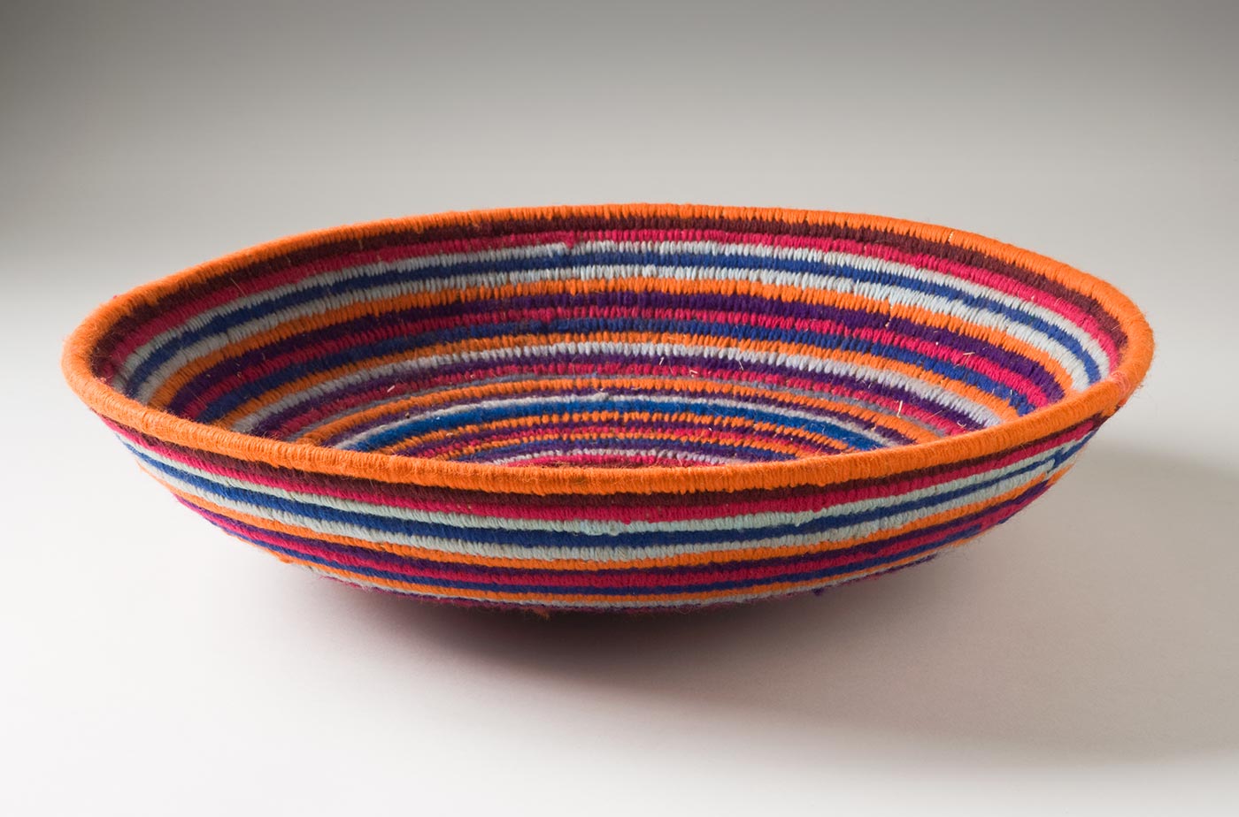 A multi-coloured circular coiled bowl-shaped basket made of yarn and plant fibre. The centre of the basket is in orange yarn followed by horizontal stripes of yarn in dark pink, purple, grey, blue, brown and orange. - click to view larger image