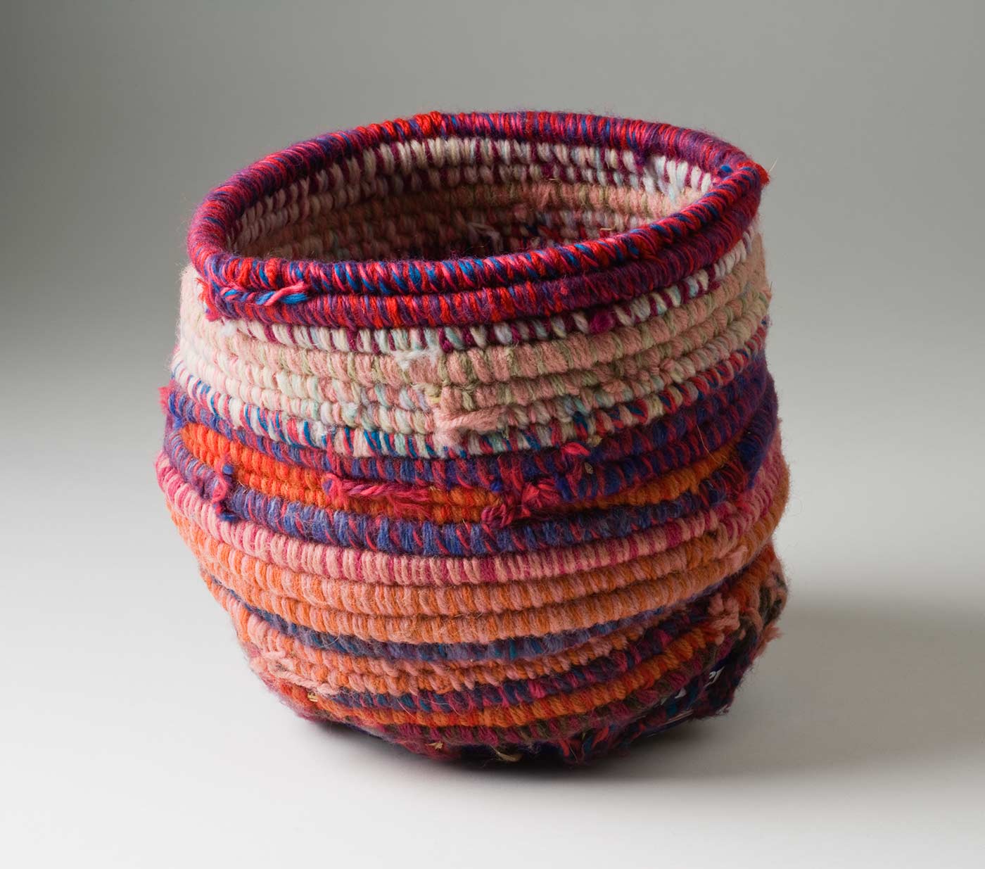 A cylindrical coiled multicoloured yarn and plant fibre basket with a metal base. The light and dark pink coiled yarn is mixed with colours of blue, orange, red and purple. The base of the basket is a blue and white coloured metal tin with burn marks to the bottom. Yarn is used to attach the base to the top section through holes that have been punched in the tin. - click to view larger image
