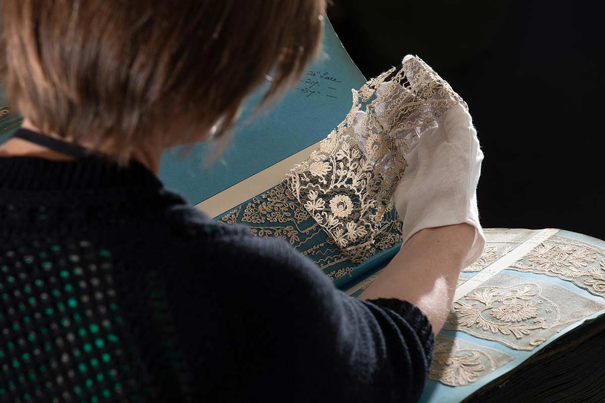 Colour photograph of a woman wearing conservation gloves and examining lace samples from a large sample book. - click to view larger image
