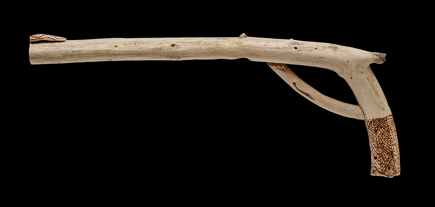 An artwork, in the shape of a pistol made from driftwood, with poker work design featured at the lower end of the handle. - click to view larger image