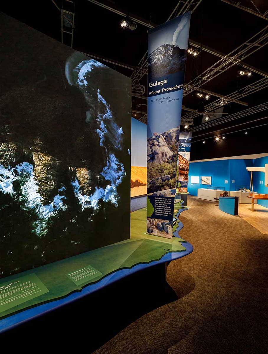 Interior view of a museum exhibition with a large image of a coastline in the foreground and a section featuring parts of a ship's interior in the distance. - click to view larger image