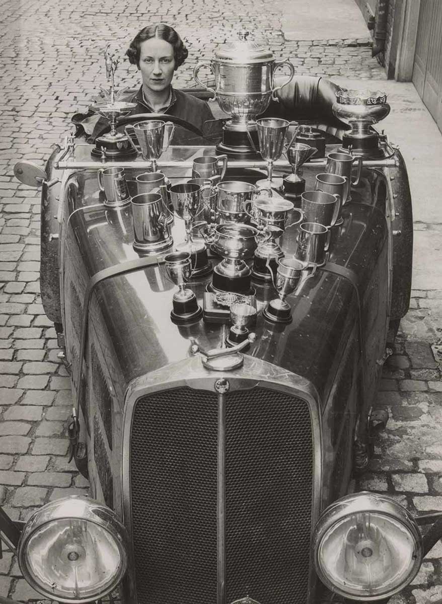 Black and white portrait-oriented photograph, showing Joan Richmond sitting in the driver’s seat of her race-car, with a large number of trophies placed on the car’s bonnet. - click to view larger image