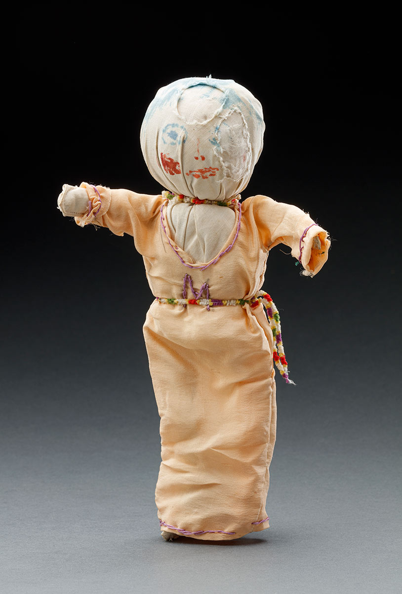 A discoloured white cotton fabric doll with the hair and facial features drawn on in ink. Her outfit consists of a peach dress with purple thread embroidery around the 'v' neckline, sleeve cuffs and hem. An embroidered letter 'M' is sewn to the centre front of the dress and the garment fastens at the back by a metal hook and eye closure. Accessories include a fuzzy multicoloured cord belt and choker. - click to view larger image