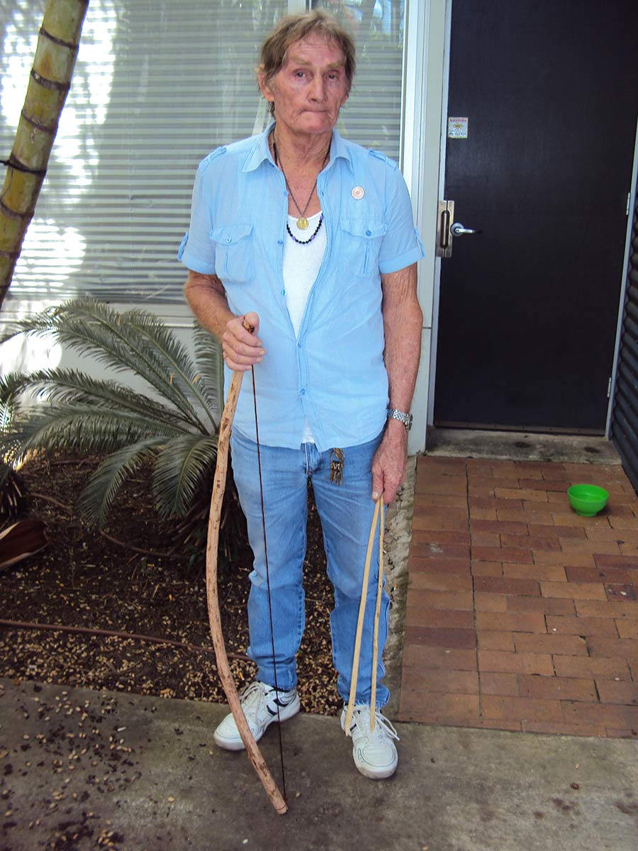 An elderly man in casual clothing is holding a crude looking bow and arrows. - click to view larger image