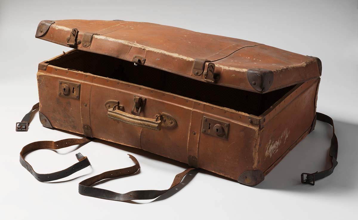 A brown suitcase featuring a wooden handle attached to the top by metal clips, leather corner protectors and metal clasps a a pair of brown and black leather and metal suitcase straps. - click to view larger image