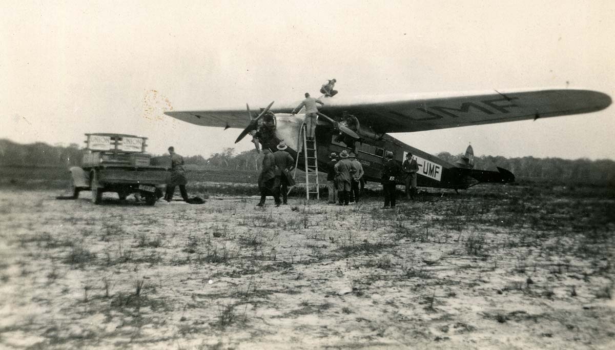 Black and white photograph of a small aircraft on the ground. People are gathered around and on top of it.