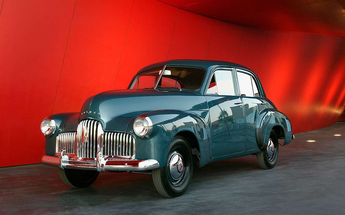 Four door, blue-gunmetal grey sedan with chrome-plated radiator grille, bumper bars, and hub caps. The red entryway to the National Museum forms a backdrop. - click to view larger image