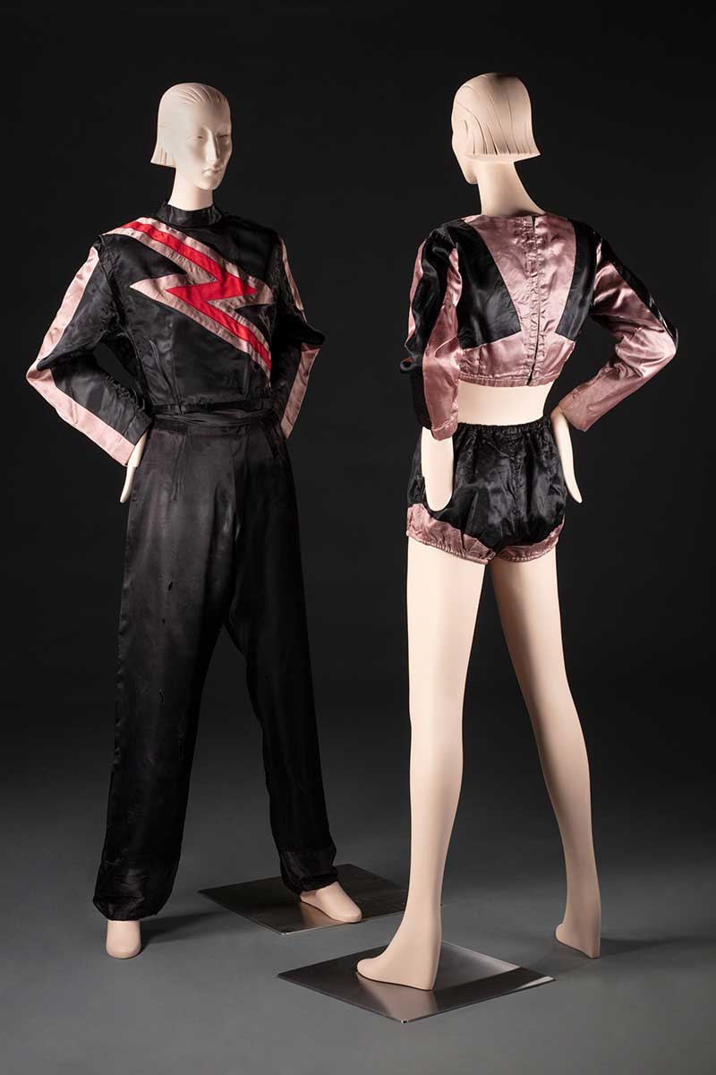 Two mannequins displaying costumes. One is a full length black and salmon pink jumpsuit with a lightning bolt motif, the other a black and salmon pink costume with crop top and bloomers. - click to view larger image