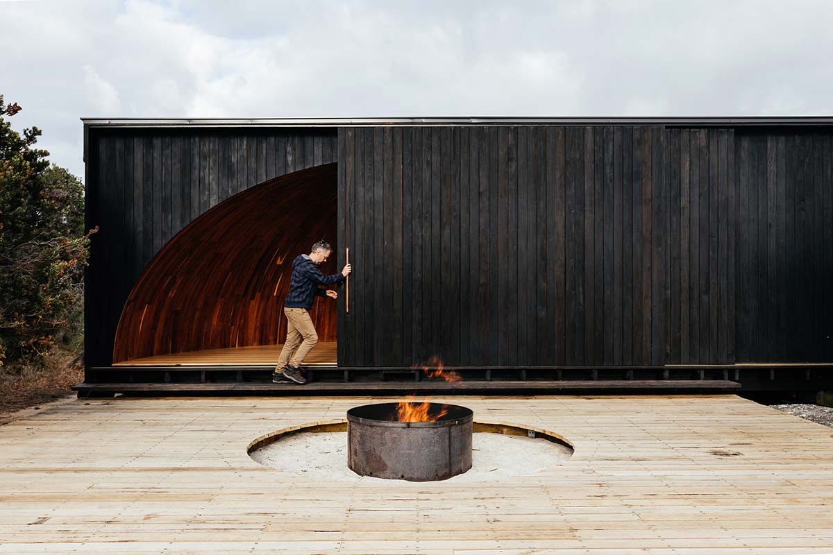 Promotional photograph featuring a man pushing a large sliding door open of a black wood cladded building. There is a deck area with a fire pit in the foreground. - click to view larger image