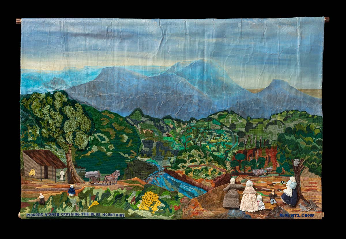 The top of the mural is painted on canvas and depicts a mountain range against a blue sky. The bottom of the mural is a mix of embroidery and tapestry. It depicts green rolling hills in the background with various people performing different tasks in the foreground, and a building on the left-hand side. The people are accompanied by animals including horses and kangaroos, and a koala in a tree on the right. Embroidered along the bottom are the words ‘PIONEER WOMEN CROSSING THE BLUE MOUNTAINS’ and ‘BLUE MTS. GROUP’.