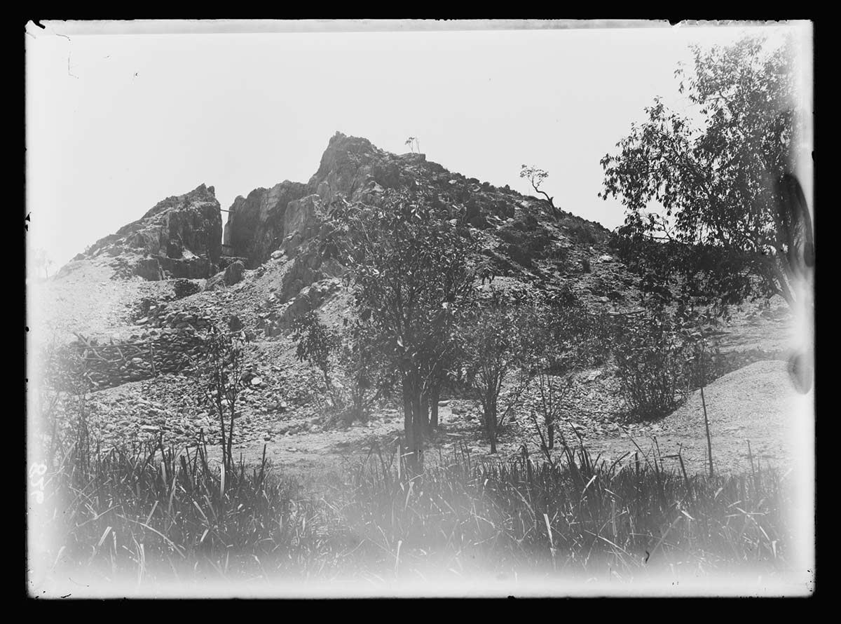 Open-cut mine formation, Daly River Copper Mine, Northern Territory probably 1905. The open-cut mine is on the left side of a hill in the background. It's seen as a jagged gash in the hillside; there is loose rock on the eroded slope in front of the mine. The hill slopes toward an area of scattered trees and long grass in the middle and foreground. - click to view larger image