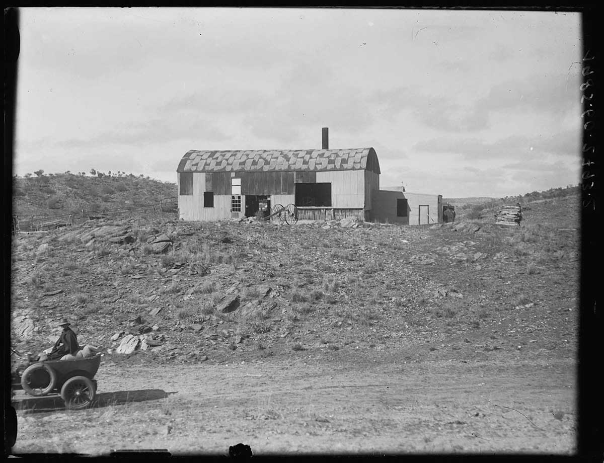 Battery building where gold-bearing rock was crushed, Arltunga, Northern Territory 1923. The building has a rectangular form and a rounded roof. It sits up on a raised section of land above an indistinct track in the foreground. A vehicle is parked in the far left foreround of the image; a man sits in the back on some baggage. The building has three openings of different sizes in the side facing the camera. A smaller building adjoins the battery at its right hand end. The battery's roofing material has weathered to a dark and light checkerboard-like pattern. A smokestack is visible toward the right end of the roof. - click to view larger image