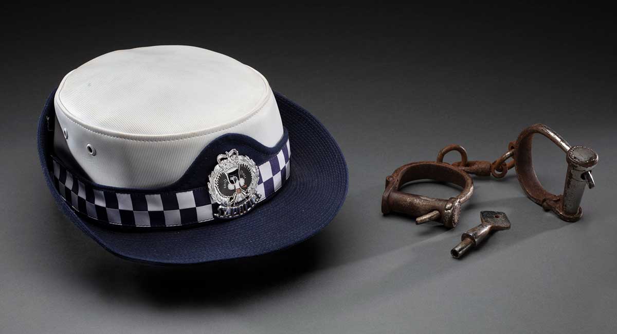 A police hat and a pair of corroded metal handcuffs with a key attached to one side on display.