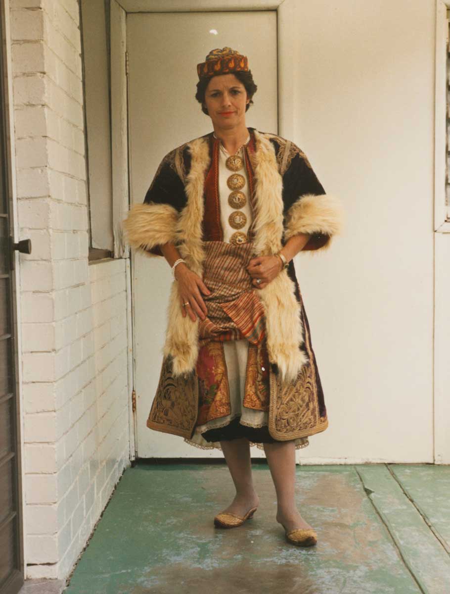 Portrait photograph of a woman with short dark hair wearing a traditional Greek garment with multiple layers. She wears a knee-length off-white chemise with long, loose sleeves. Over the chemise is a short-sleeved red vest and crimson knee-length tunic, both open at the front. The tunic is embellished with gold thread and colourful silk embroidered floral motifs. The woman wears five gold button brooches, running down the chest of the chemise, and a pair of embroidered gold slippers with a small heel. Wrapped around her waist and hips is a woven sash with fine checked pattern. Worn over the sash and tunic is a deep purple-coloured three-quarter length overcoat with cream-coloured fur trim down the front opening and hems of the sleeves. The woman wears a yellow, red and blue beanie-shaped hat, and stands facing the camera. - click to view larger image