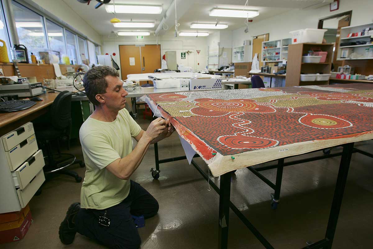 A conservator kneels to inspect edge of large canvas, with laboratory in background. - click to view larger image